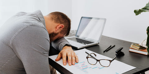 A Caucasian man wearing a checkered shirt putting his head down on a table in front of a laptop